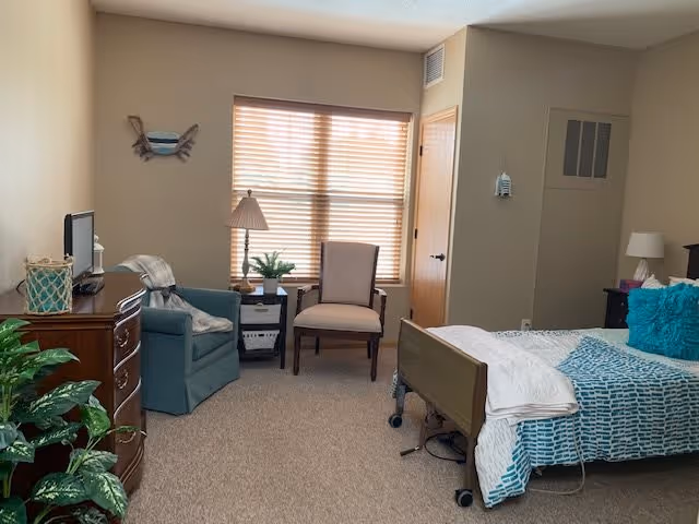 A cozy bedroom in an assisted living facility featuring a bed with blue and white bedding, a wooden dresser with a TV on top, two chairs near a window with blinds, a small side table with a lamp and plant, and a potted plant in the corner.