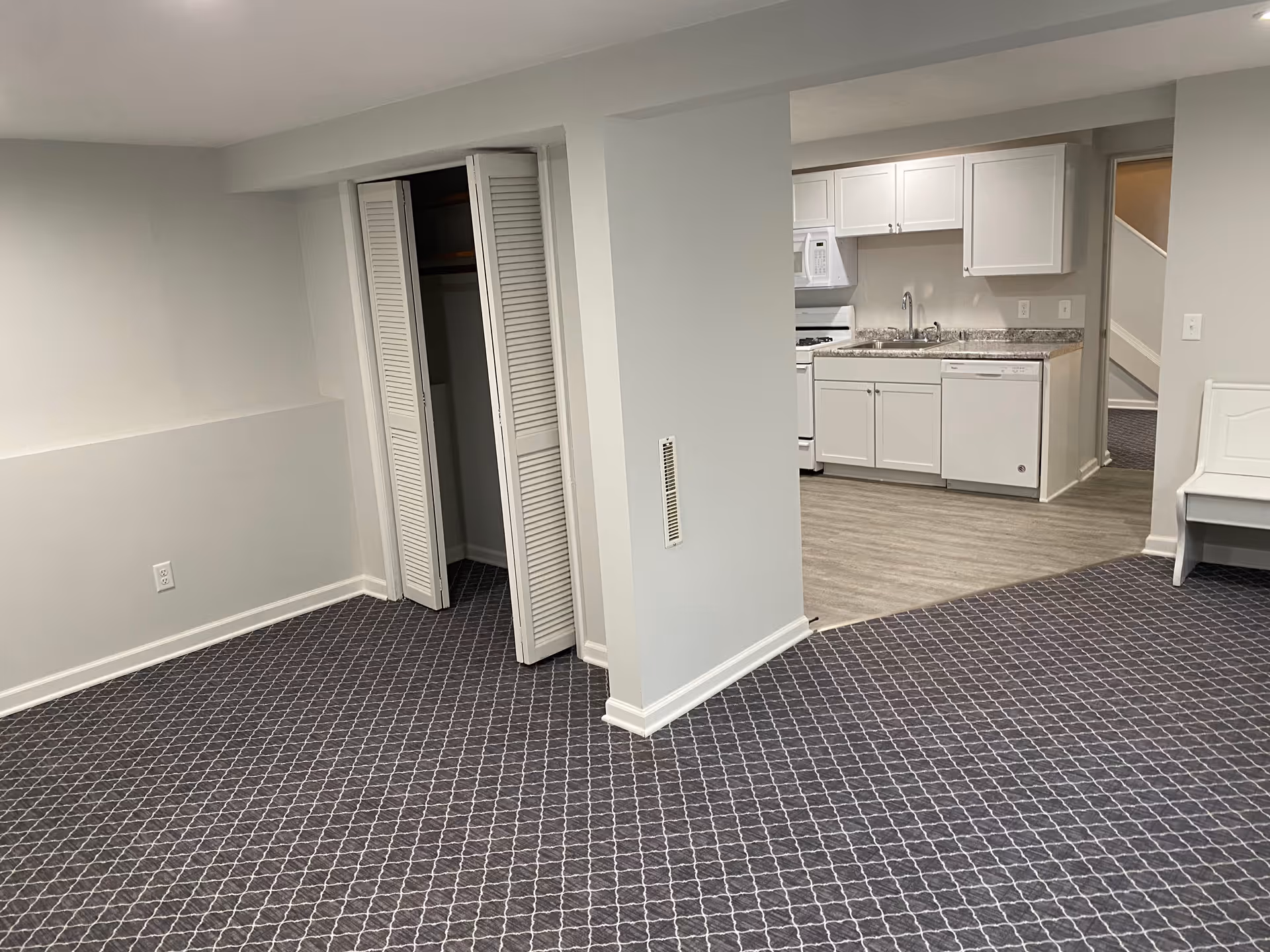 Interior view of a room with patterned carpet flooring, an open closet with white louvered doors, and a small kitchen area with white cabinets, a sink, a microwave, and a dishwasher. There is also a white bench against the wall and a staircase visible in the background.