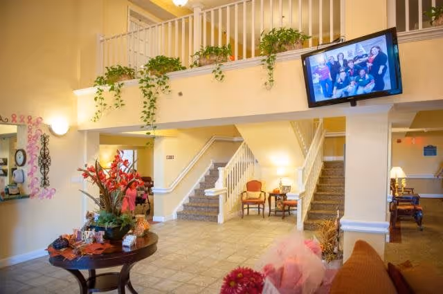 Interior view of a senior living facility lobby with a staircase leading to an upper floor. The area features a round table with a floral arrangement, potted plants on the railing above, chairs arranged near the stairs, and a wall-mounted TV displaying a group photo. The walls are painted light yellow and the floor is tiled.