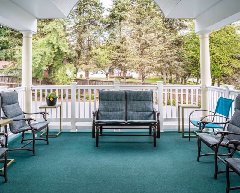 Covered outdoor porch area with green carpet flooring, white railing, and several cushioned chairs and a loveseat arranged around small side tables. Trees and a wooden fence are visible in the background.