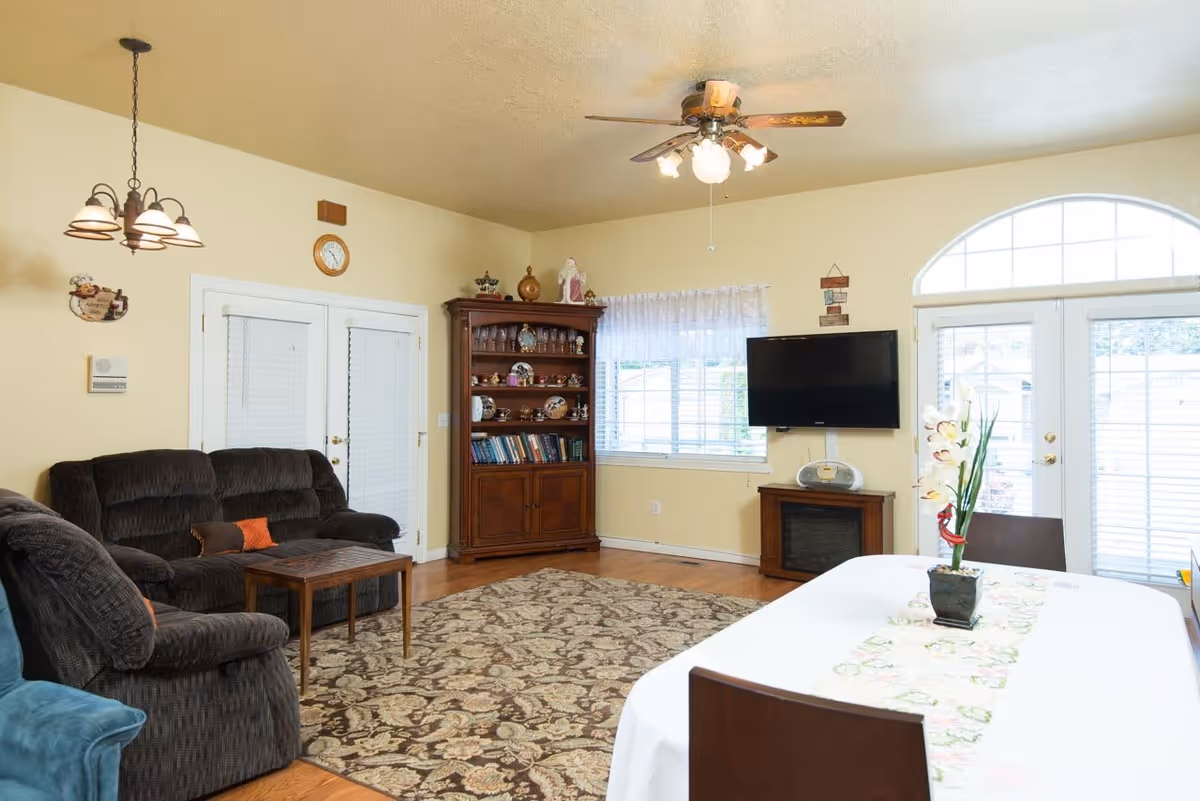 A cozy living room with a dark brown sofa and armchair, a wooden coffee table, a patterned area rug, a wooden cabinet filled with decorative items and books, a wall-mounted flat-screen TV above a small cabinet, a window with white blinds and sheer curtains, a ceiling fan with lights, and a dining table with a white tablecloth and a floral centerpiece. French doors with blinds lead outside.