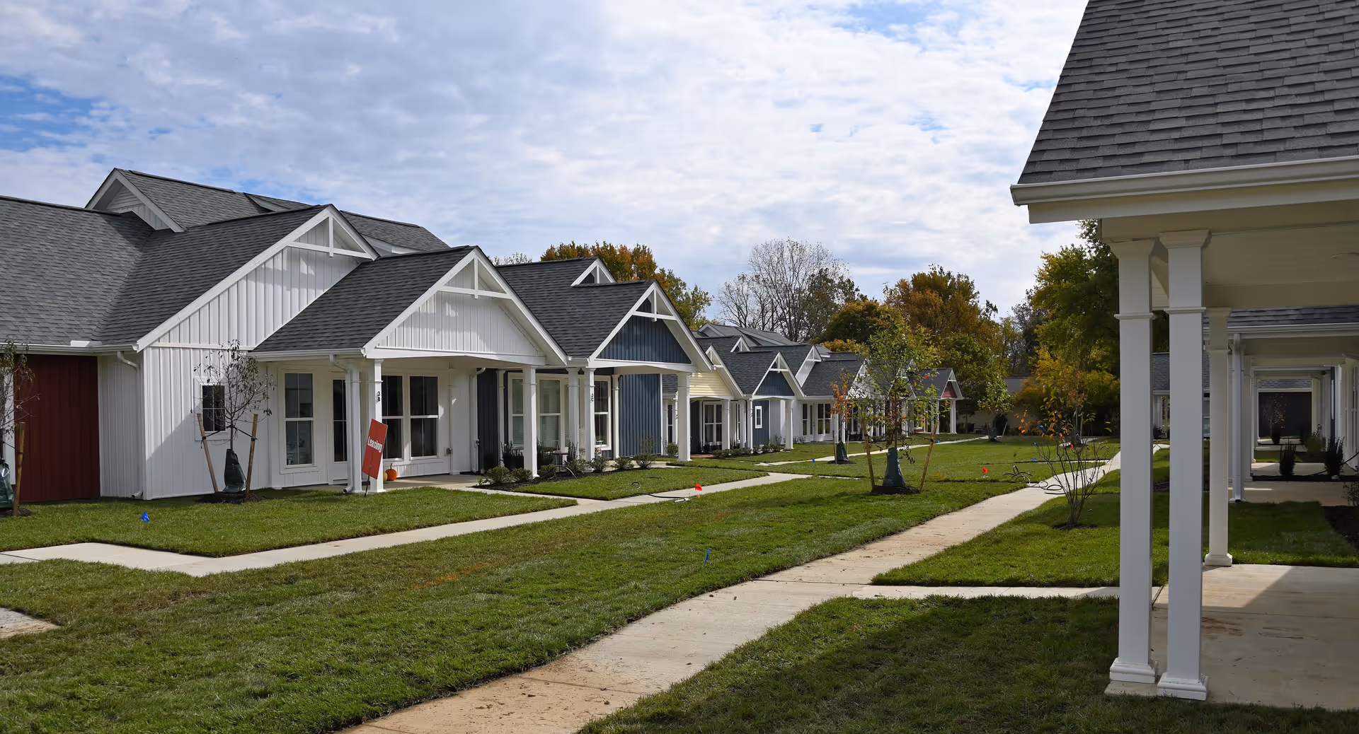A row of single-story residential buildings with pitched roofs and front porches, set along paved walkways with green lawns and young trees under a partly cloudy sky.