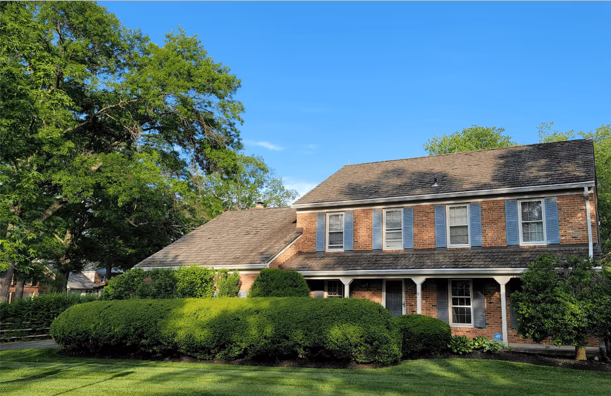 A two-story brick house with blue shutters and a shingled roof surrounded by green bushes and trees under a clear blue sky.
