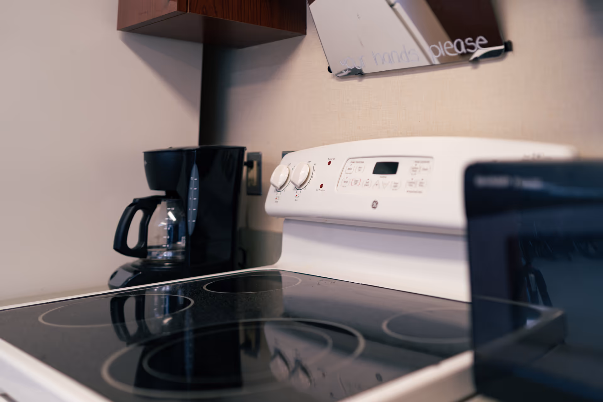 Close-up view of a kitchen stove with a smooth black glass cooktop and white control panel. A black coffee maker with a glass pot is placed on the counter next to the stove. A mirror or reflective surface is partially visible above the stove with faint text on it.