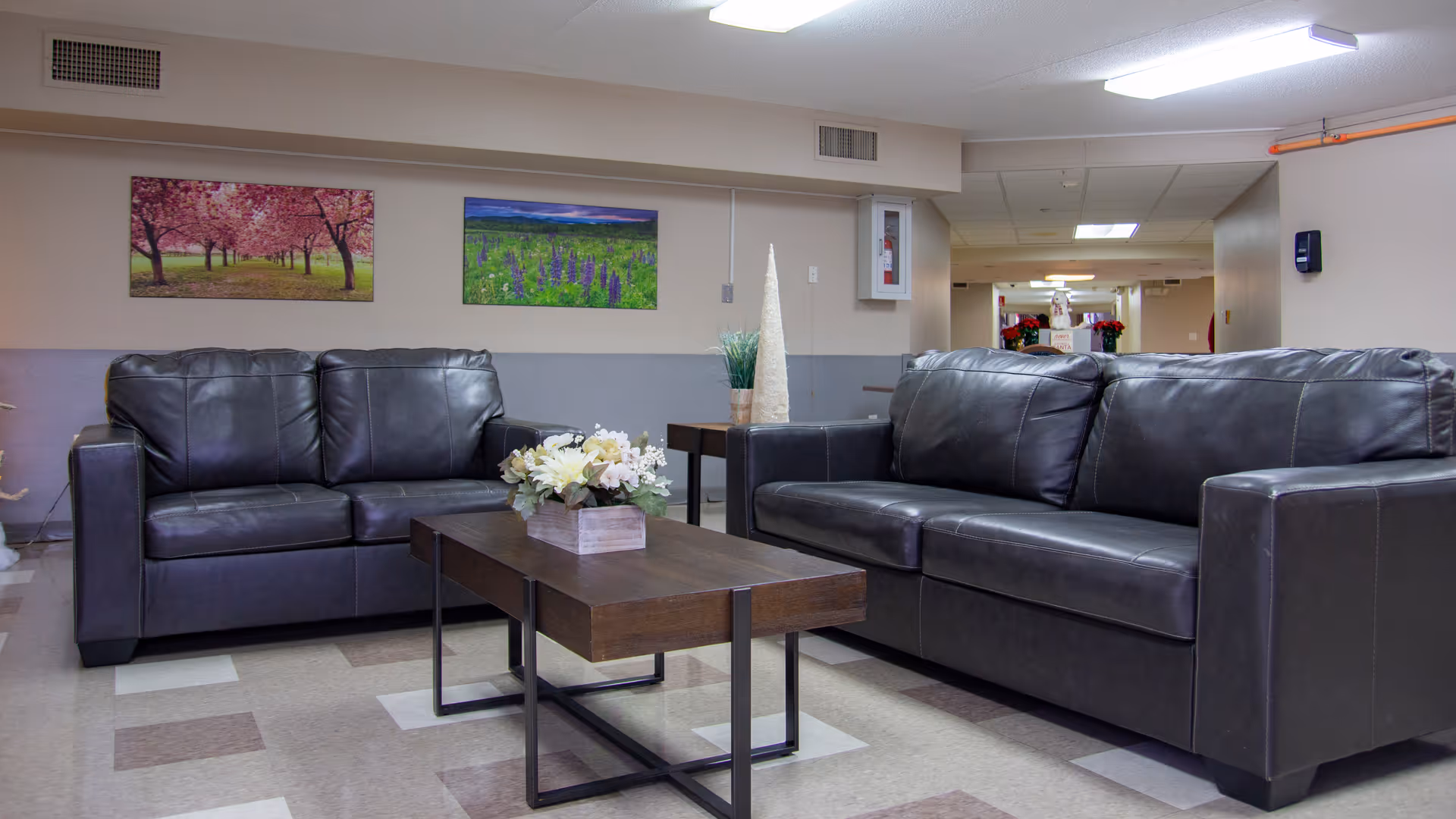 A seating area in a senior living facility with two dark leather sofas facing each other and a wooden coffee table with a flower arrangement in the center. The walls are painted beige and gray, decorated with two landscape paintings. The background shows a hallway with ceiling lights and some holiday decorations.
