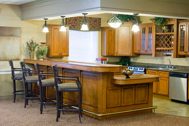 A warm and inviting kitchen area with wooden cabinetry, a granite countertop, and a breakfast bar with four high chairs. The kitchen features a stove, dishwasher, and a wine rack. Pendant lights hang above the bar, and there is a window with blinds behind the counter.