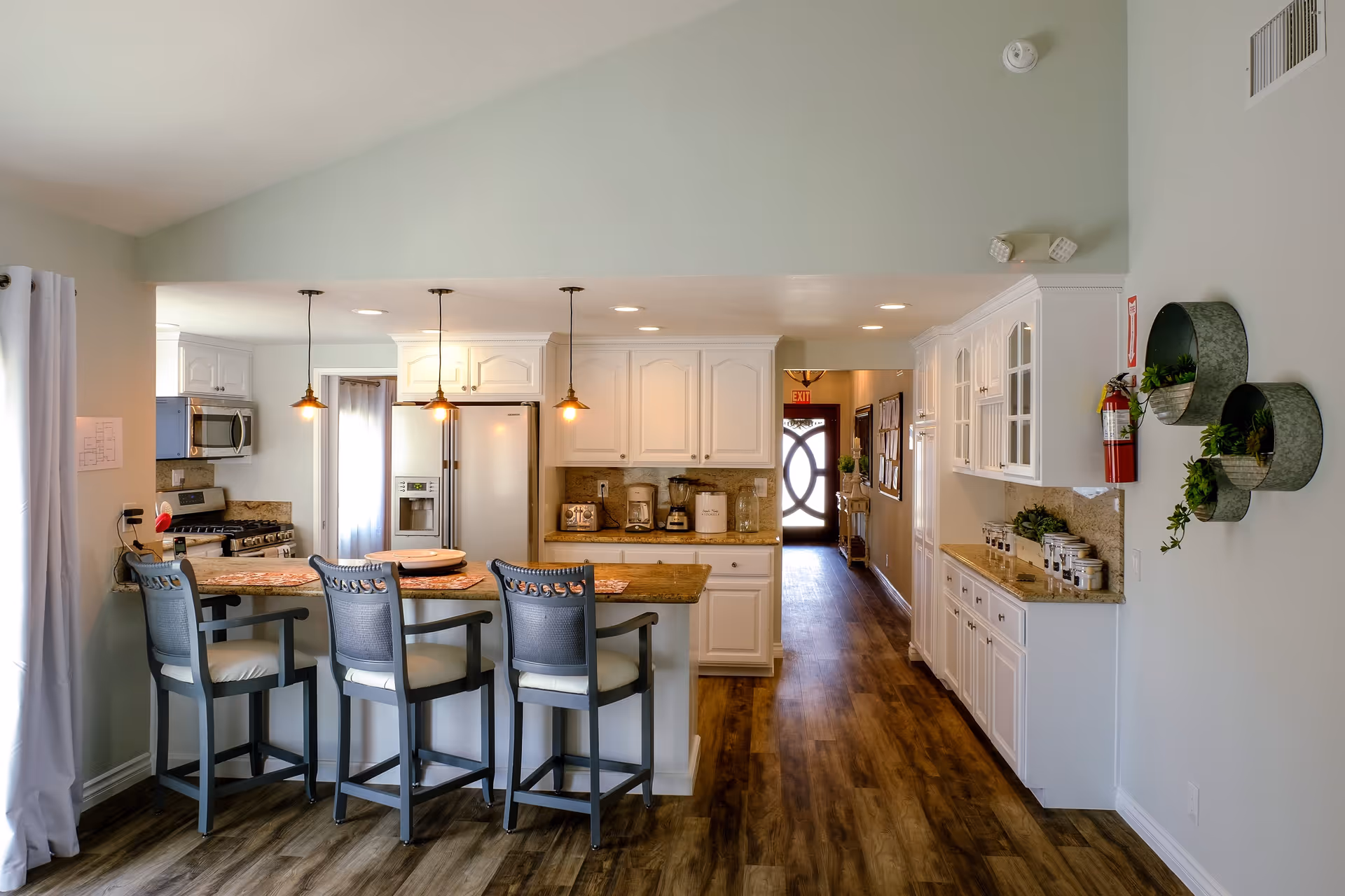 A bright kitchen area with white cabinets, granite countertops, and wooden flooring. There is a kitchen island with three high chairs, pendant lights hanging above, and various kitchen appliances including a refrigerator, microwave, toaster, coffee maker, and blender. On the right wall, there are decorative round metal wall planters with green plants and a fire extinguisher mounted. A hallway with wooden flooring leads to a door with a circular window design.