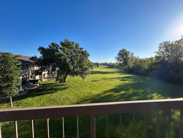 View from a balcony overlooking a large grassy area with several trees and a building on the left side under a clear blue sky.