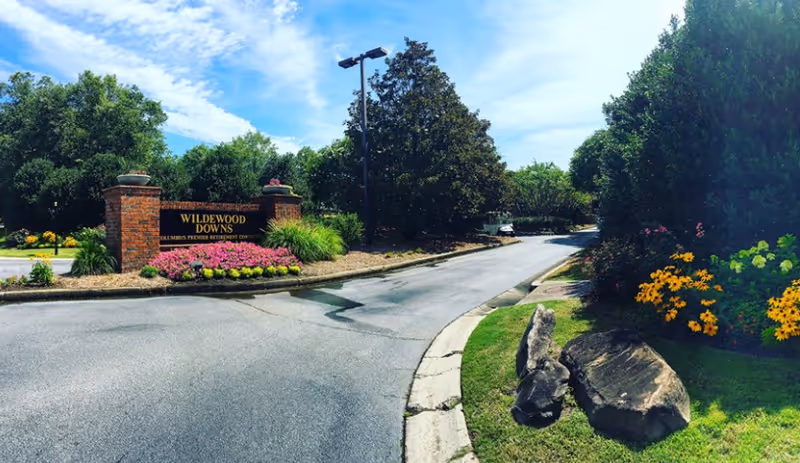 Entrance driveway and landscaped brick sign reading "Wildewood Downs" surrounded by flowers, rocks, and trees.
