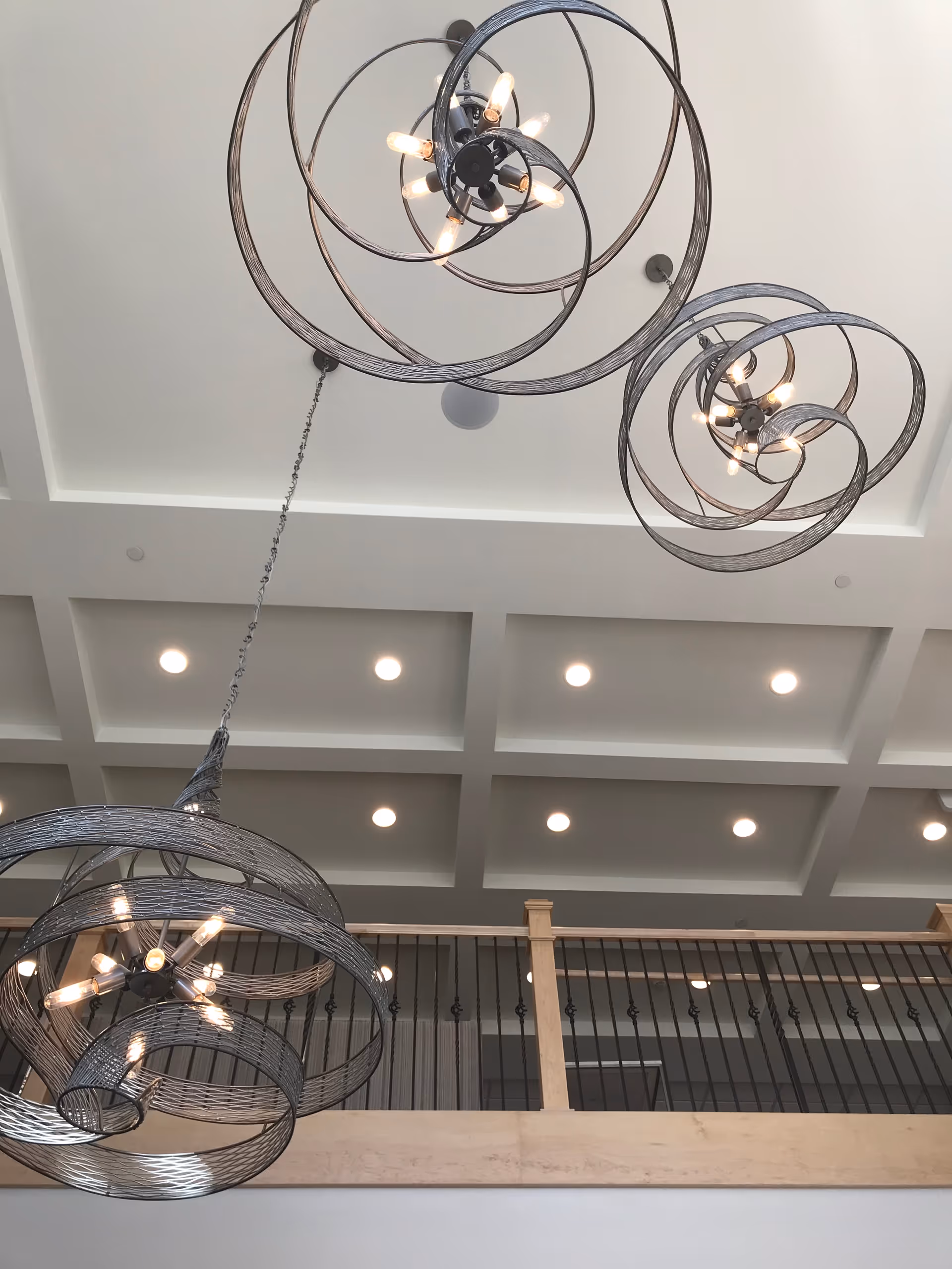 View of a high ceiling with modern spiral metal chandeliers and recessed lighting, above a wooden balcony railing with black metal balusters.