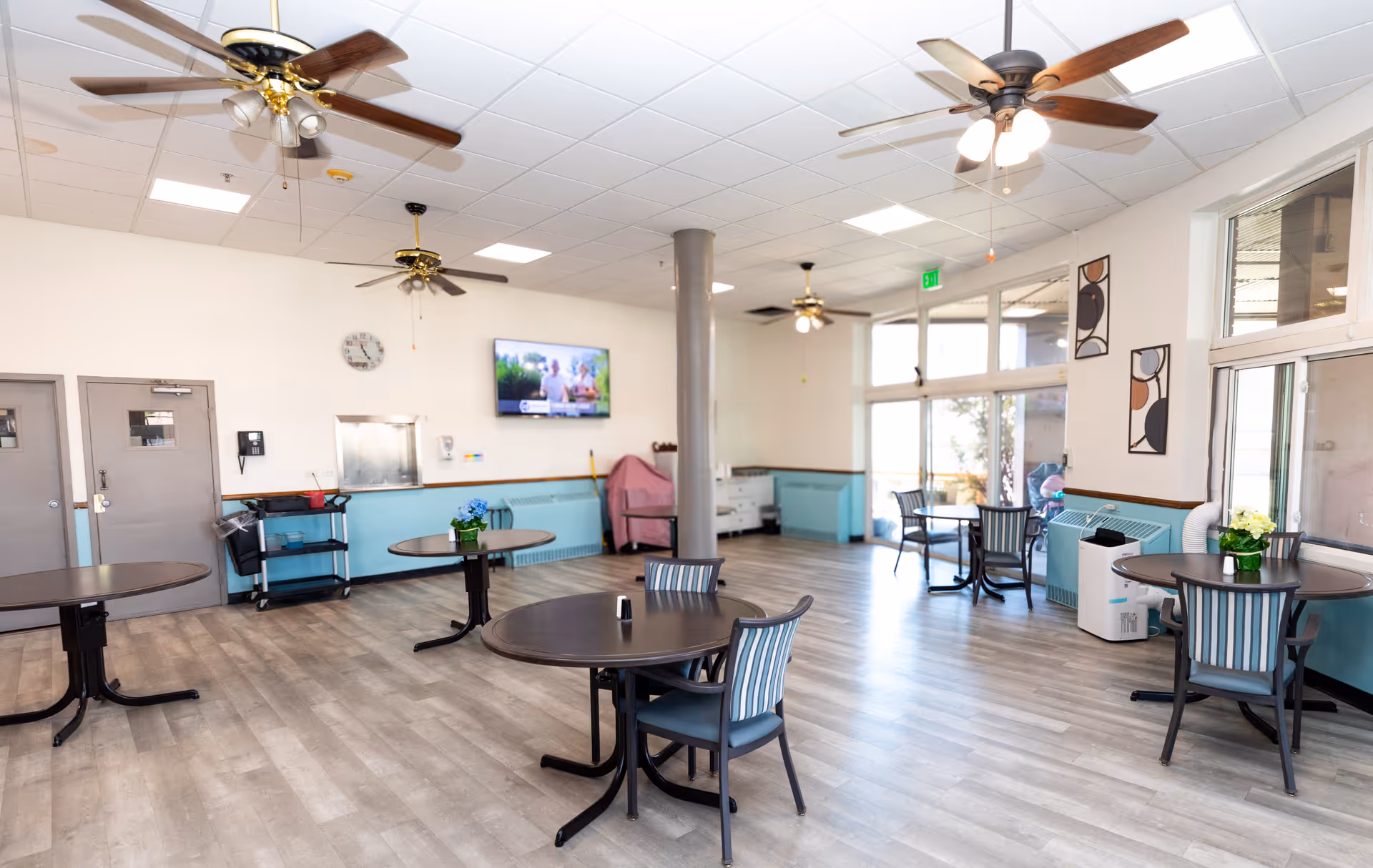 A bright and spacious dining area with several round tables and chairs arranged throughout the room. The room features light wood flooring, ceiling fans with lights, large windows allowing natural light, and a wall-mounted television. There are decorative plants on the tables and abstract artwork on the walls.