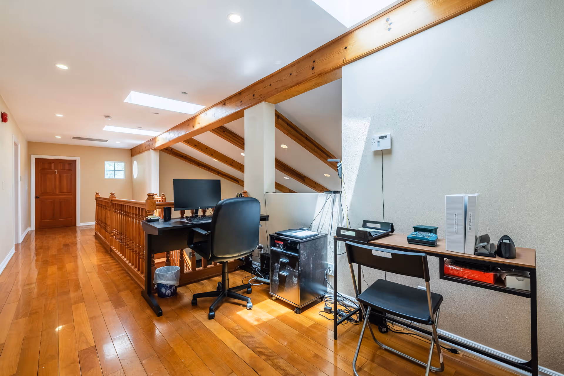 A bright hallway with wooden flooring and exposed wooden beams on the ceiling. There is a black office chair and desk with a computer monitor, printer, and other office supplies. Another small desk with a folding chair and office items is against the wall. The hallway has a wooden railing and doors at the end.