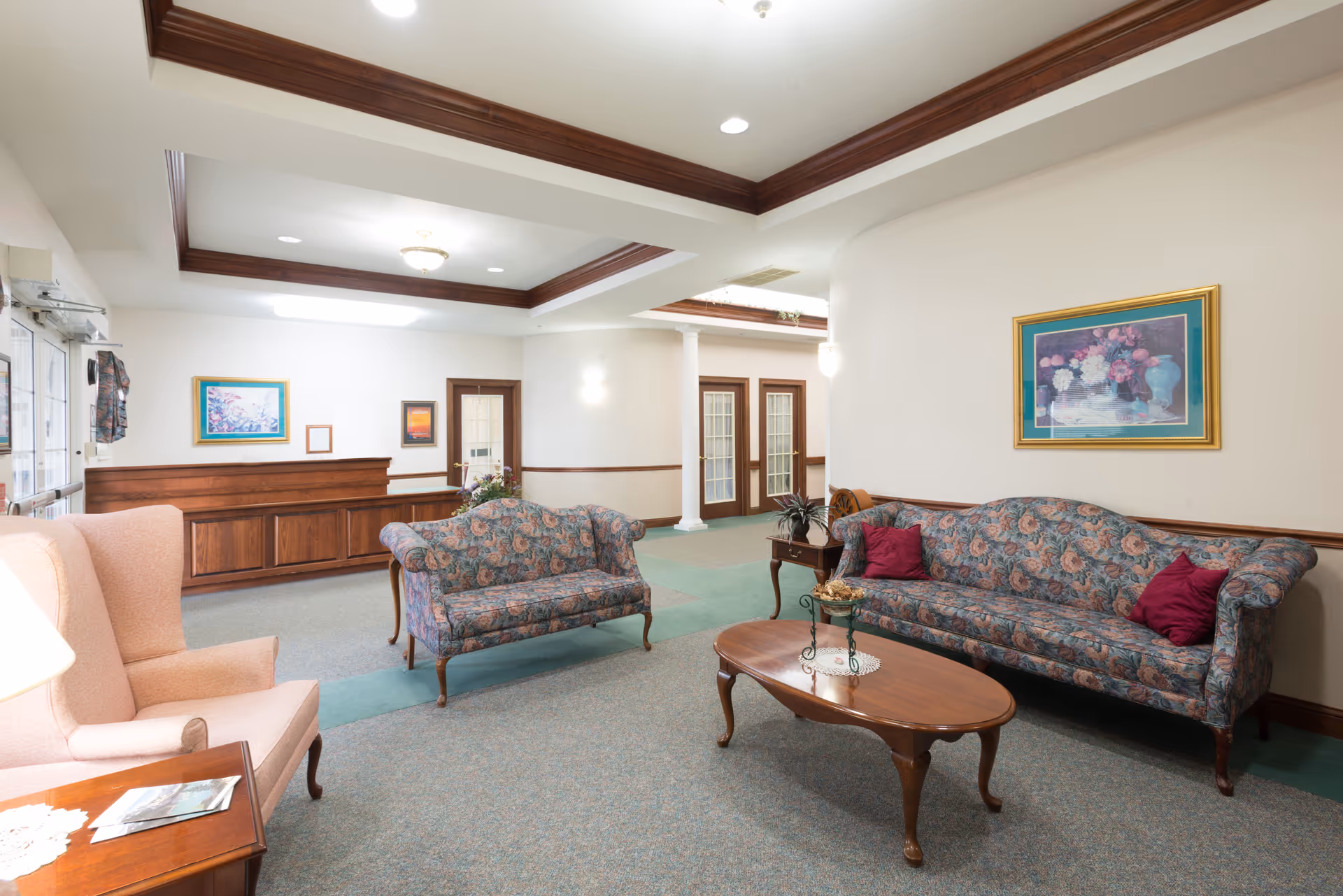 A well-lit living room area in Orchard Grove featuring floral patterned sofas with red cushions, a wooden coffee table with a decorative centerpiece, a pink armchair, framed floral artwork on the walls, and wooden trim accents on the ceiling and walls.