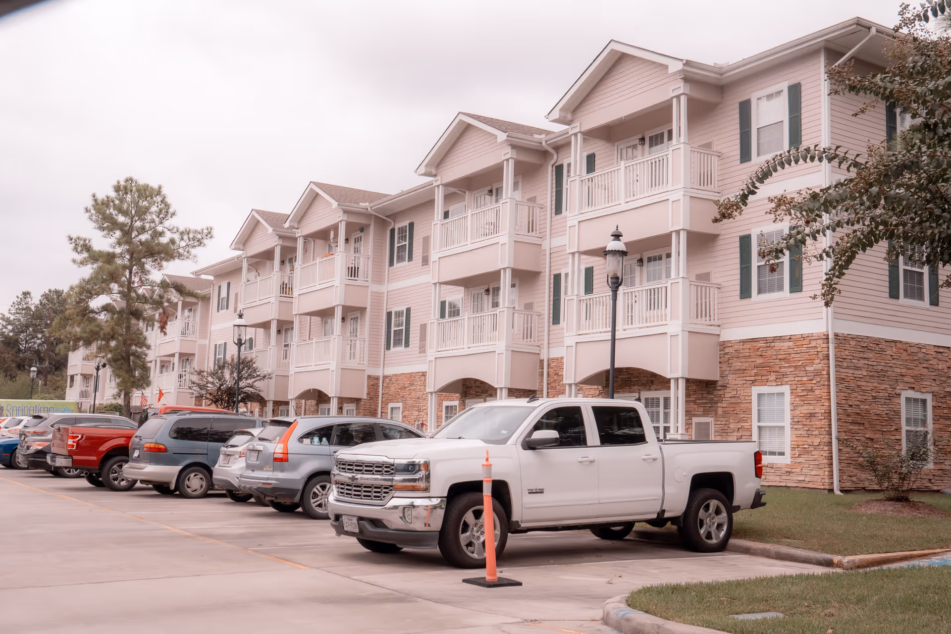 Three-story pale-pink apartment-style building with balconies and a parking lot of cars, including a white pickup in the foreground.