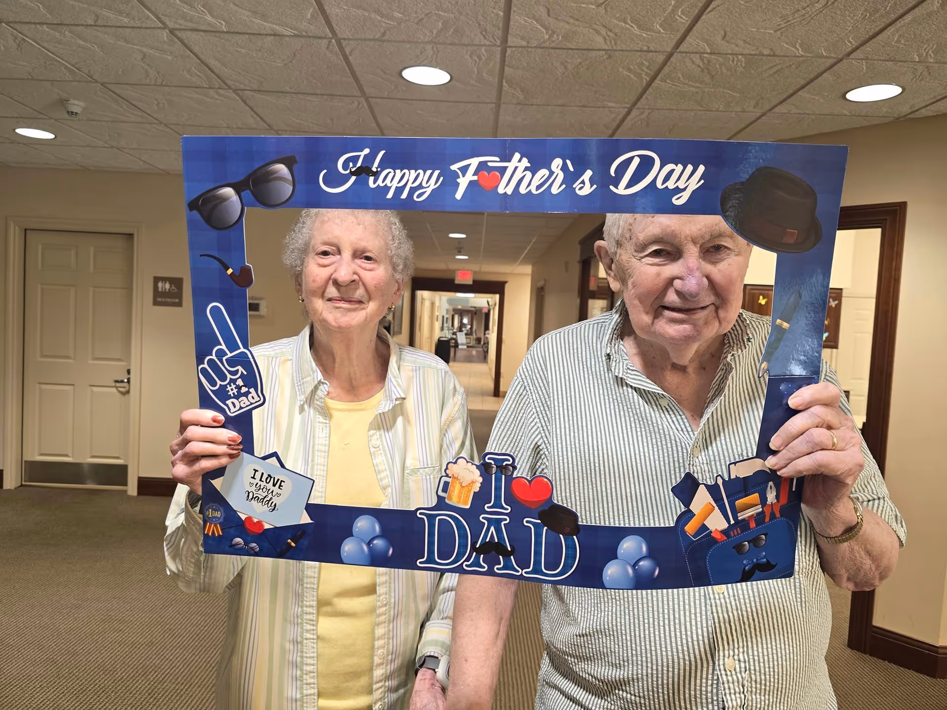 An elderly woman and an elderly man standing indoors in a hallway, holding a large blue photo frame that says 'Happy Father's Day' with various Father's Day themed decorations including sunglasses, a pipe, a foam finger, and tools. Both are smiling and looking at the camera.