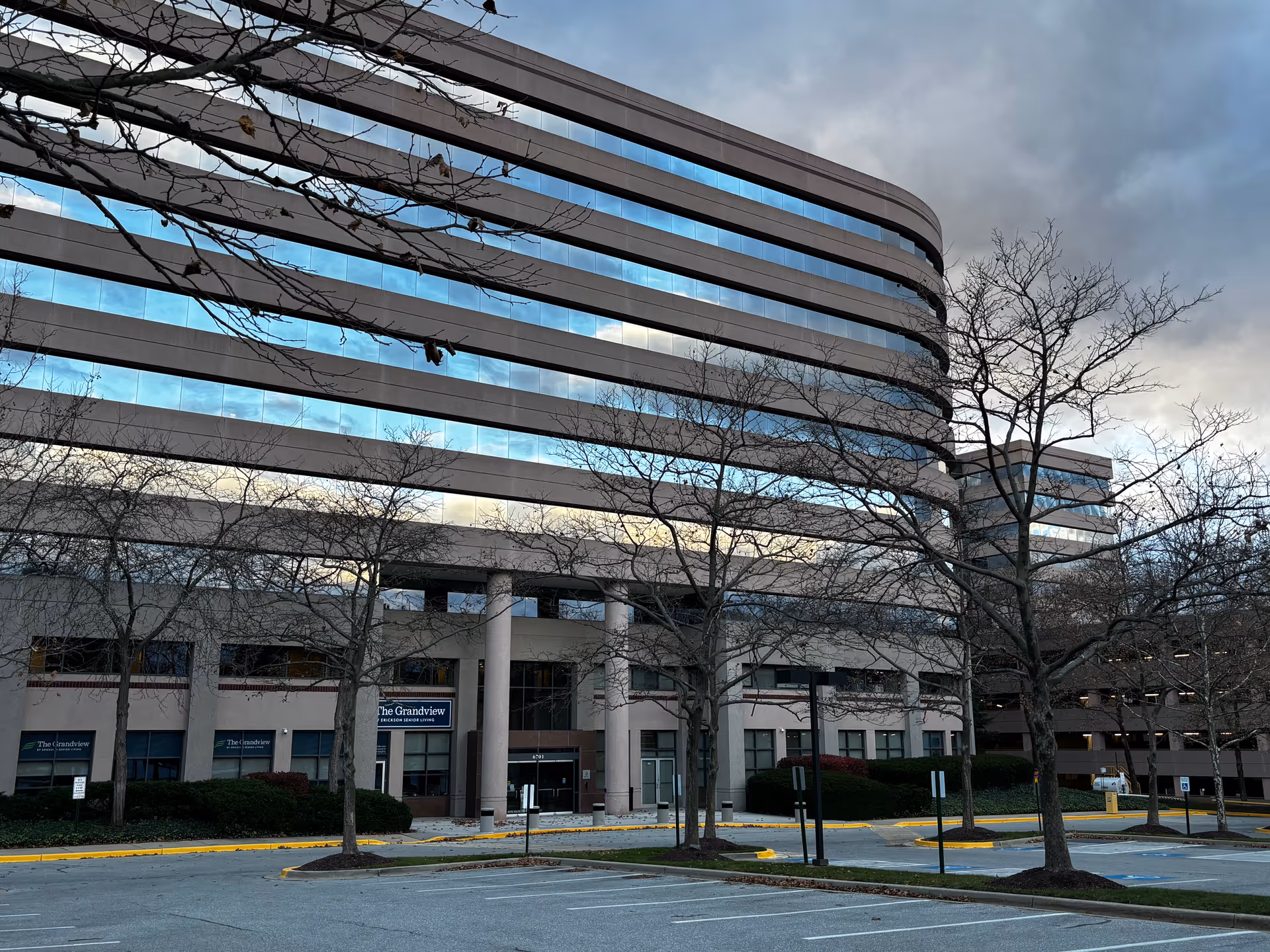 Modern multi-story office building with curved reflective windows and leafless trees in front, showing a sign for The Grandview Sales Center.