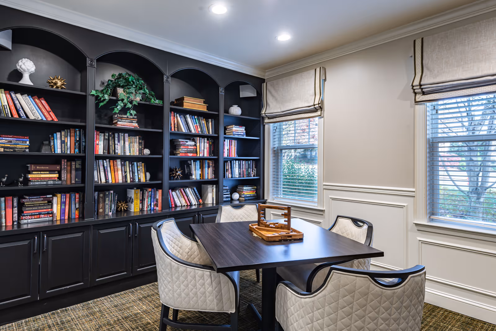 A cozy room with a dark wooden table surrounded by four cushioned chairs with quilted upholstery. Behind the table is a large black bookshelf filled with books and decorative items. Two windows with beige Roman shades allow natural light to enter the room.