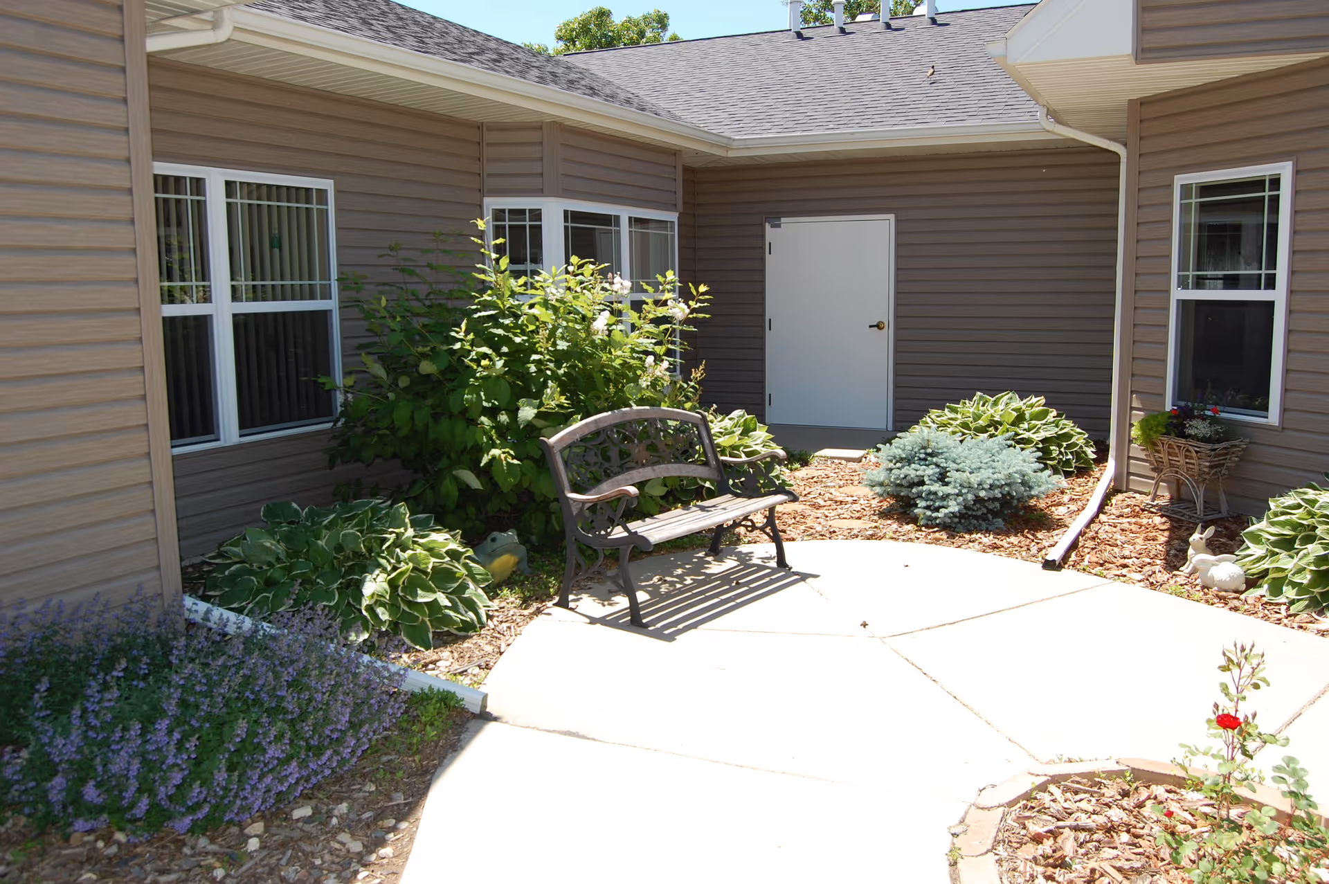 Outdoor courtyard area with a metal bench surrounded by various green plants and shrubs. The courtyard is paved with concrete and bordered by a building with beige siding and several windows. There is a white door on the building wall and decorative garden elements including small statues and a flower basket.