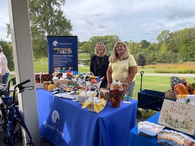 Two women standing behind a table covered with a blue Plymouth Terrace tablecloth, displaying various items including baskets, flowers, and brochures. Behind them is a blue informational banner about assisted living perks. The setting is outdoors with green grass, trees, and a cloudy sky in the background.