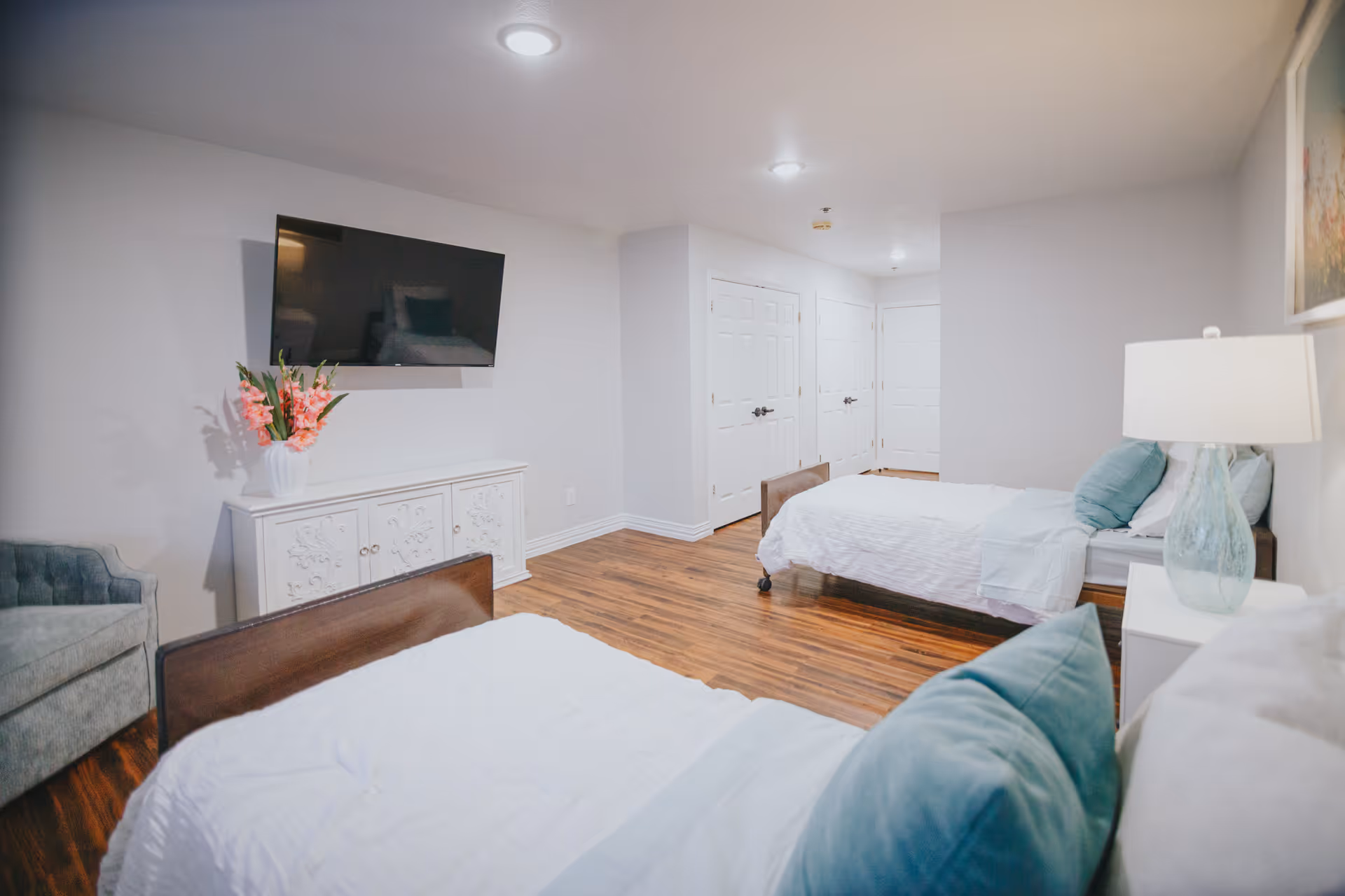 Bright, modern bedroom with two twin beds, a wall-mounted TV, dresser with flowers, and hardwood floors.