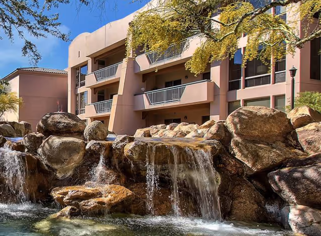 Outdoor view of a senior living facility building with balconies, surrounded by trees and a large rock waterfall feature in the foreground.