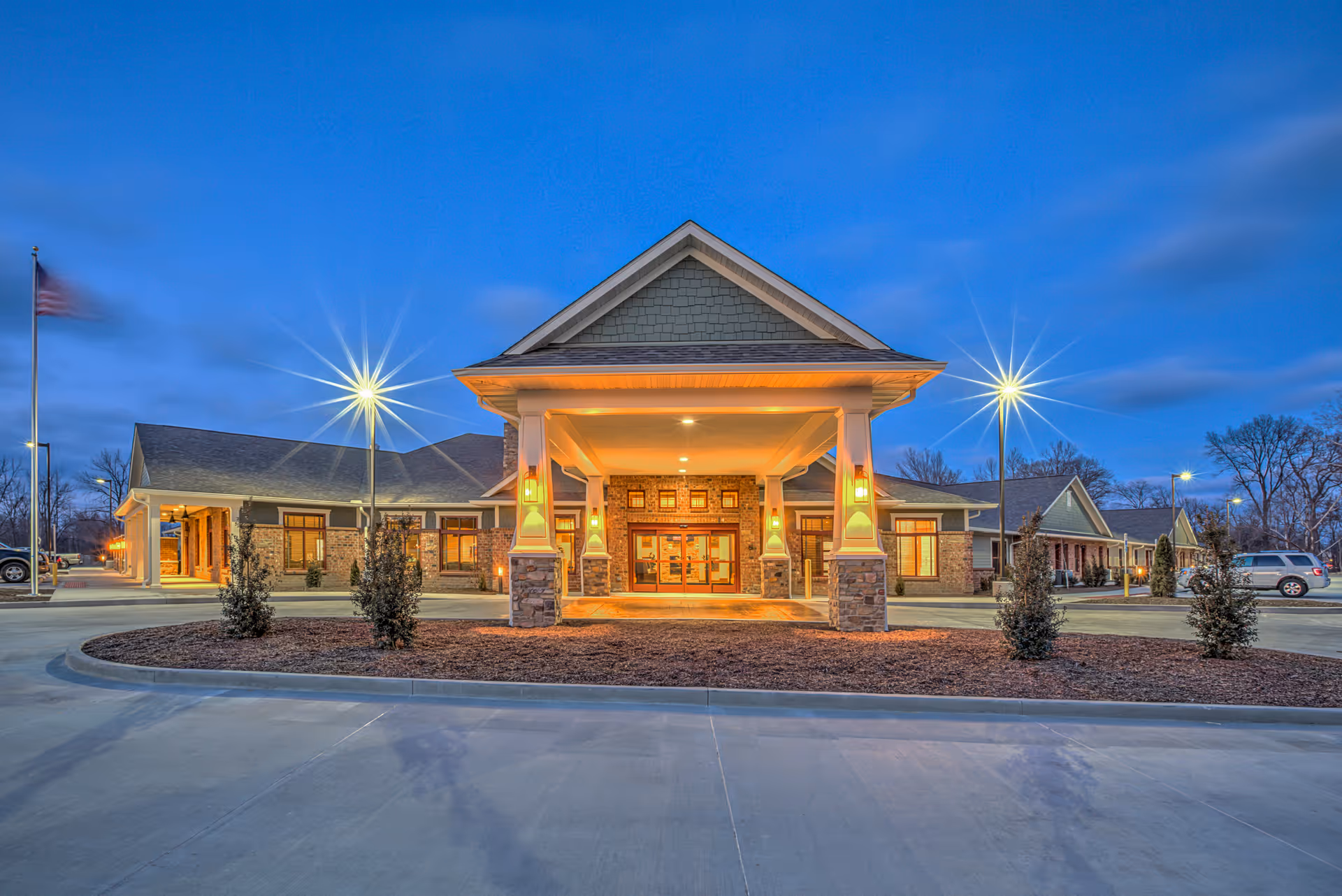 Front exterior view of Stillwater Senior Living facility at dusk with illuminated entrance, stone and brick facade, and surrounding parking area with small shrubs and an American flag on a flagpole.