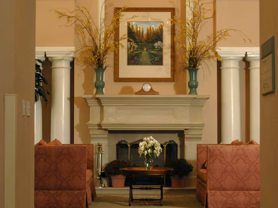 A cozy living room area with a stone fireplace in the center. On the mantel are two green vases with yellow flowers and a small clock. Above the mantel hangs a framed painting of a garden pathway. Two red patterned sofas face each other with a small wooden coffee table between them, holding a vase of white flowers. White columns flank the fireplace on both sides.