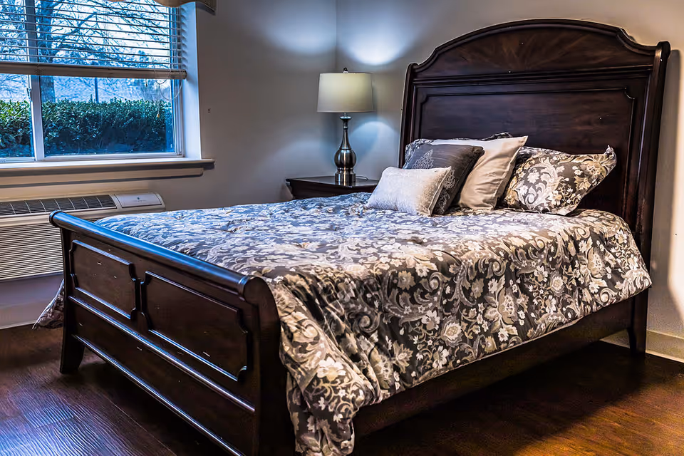 A bedroom with a dark wooden bed frame and a floral patterned bedspread. The bed has multiple pillows arranged at the head. Next to the bed is a wooden nightstand with a silver lamp. A window with blinds partially open shows greenery outside. The floor is wooden.