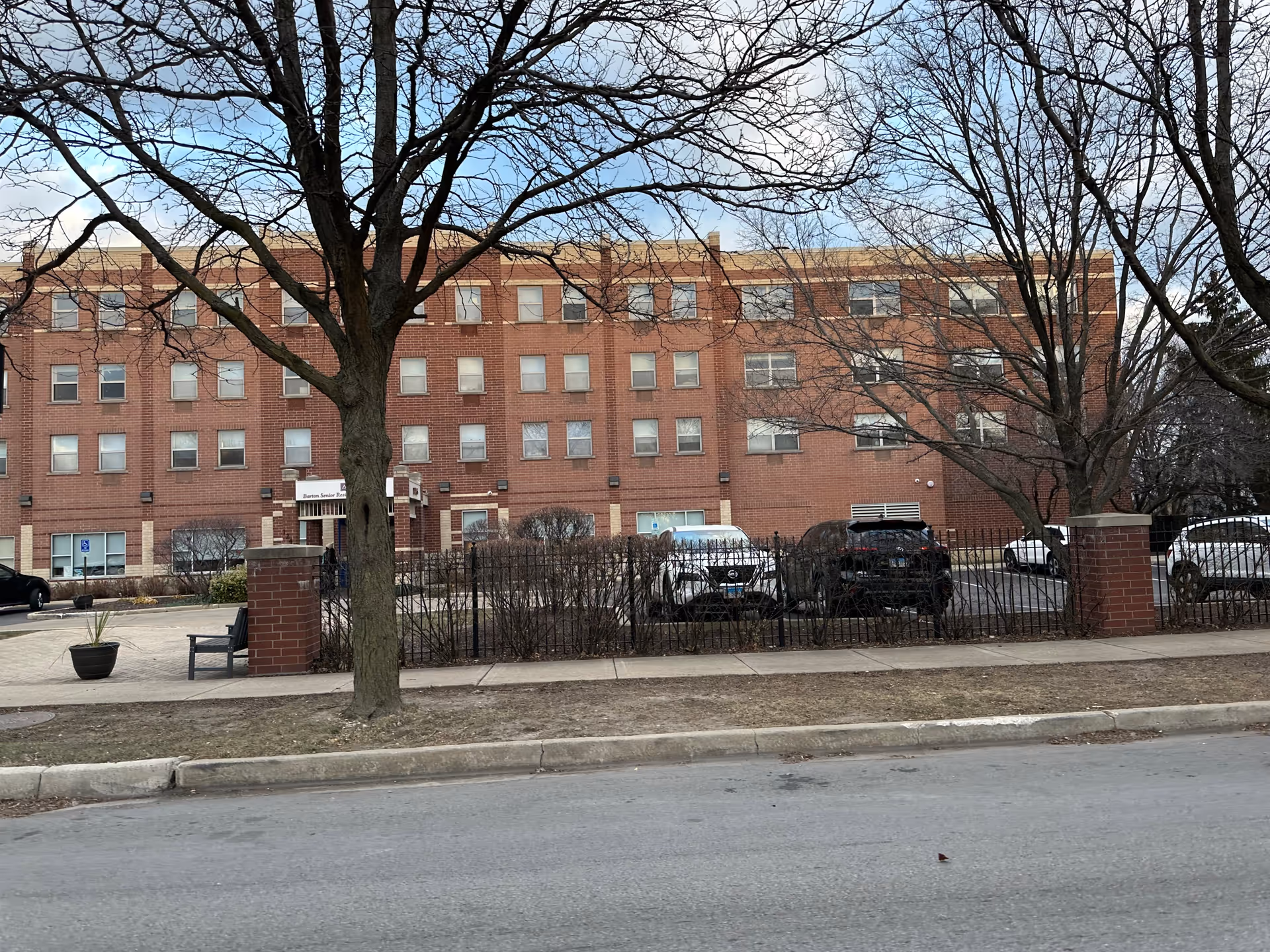 Exterior view of a three-story brick building with multiple windows, identified as Barton Senior Residences. There are leafless trees in front of the building, a sidewalk, a few parked cars, and a black metal fence with brick pillars.