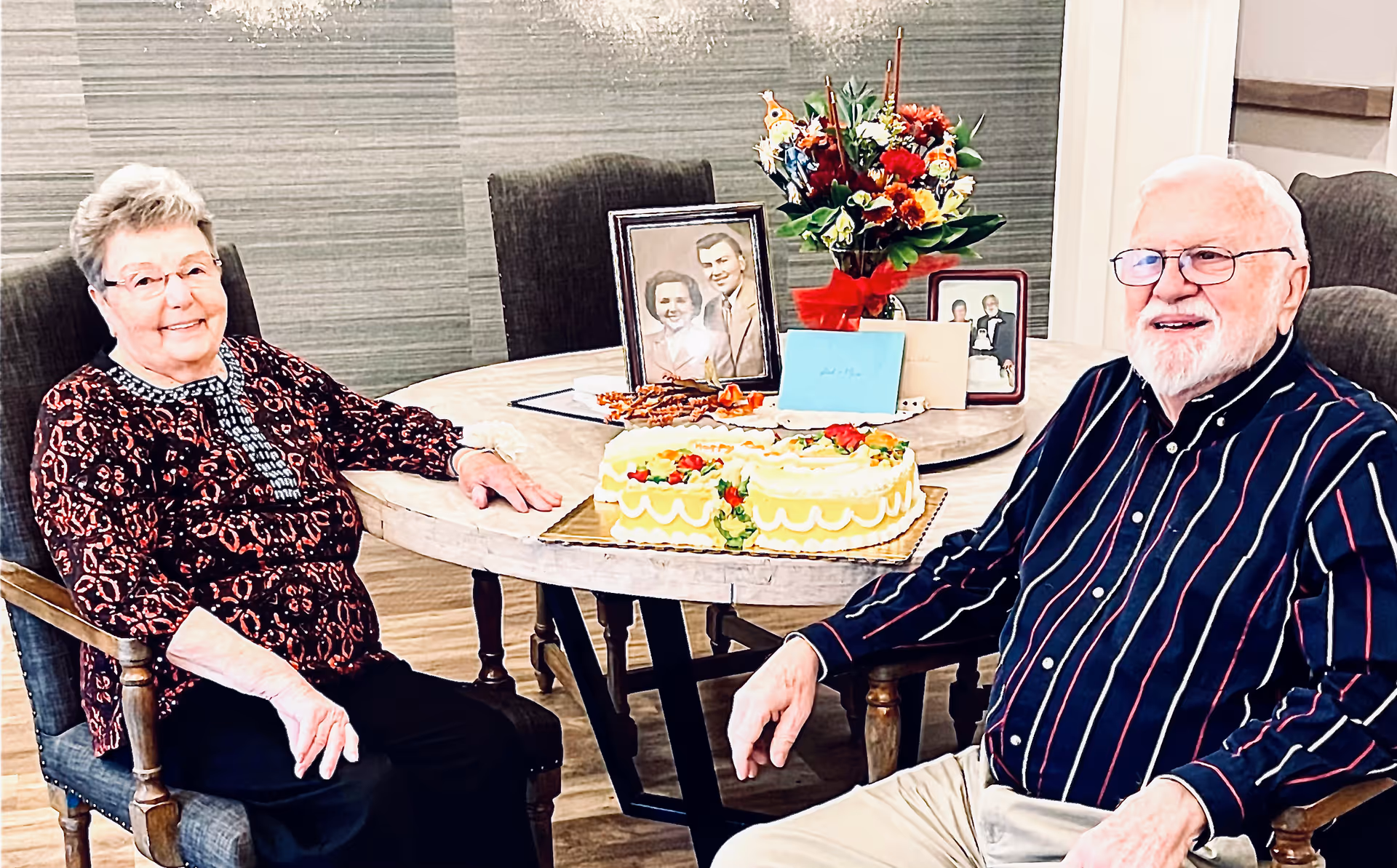 An elderly woman and an elderly man sitting at a round dining table in a well-lit room. On the table, there is a decorated cake, a bouquet of flowers, several greeting cards, and framed photographs. Both individuals are smiling and appear to be enjoying a celebratory moment.