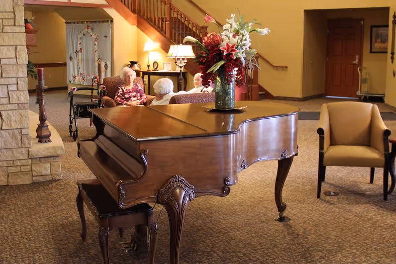 A cozy common area in a senior living facility featuring a grand piano with a floral arrangement on top, several armchairs, and elderly residents sitting and conversing near a staircase and a stone fireplace.