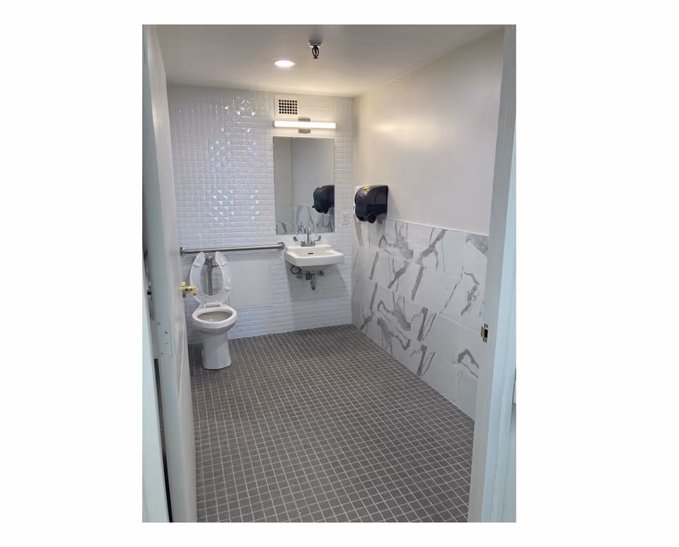 A clean, accessible bathroom with a toilet, a wall-mounted sink, a mirror above the sink, and two black paper towel dispensers on the wall. The floor is covered with small gray tiles, and the walls have white and gray marble-patterned tiles.