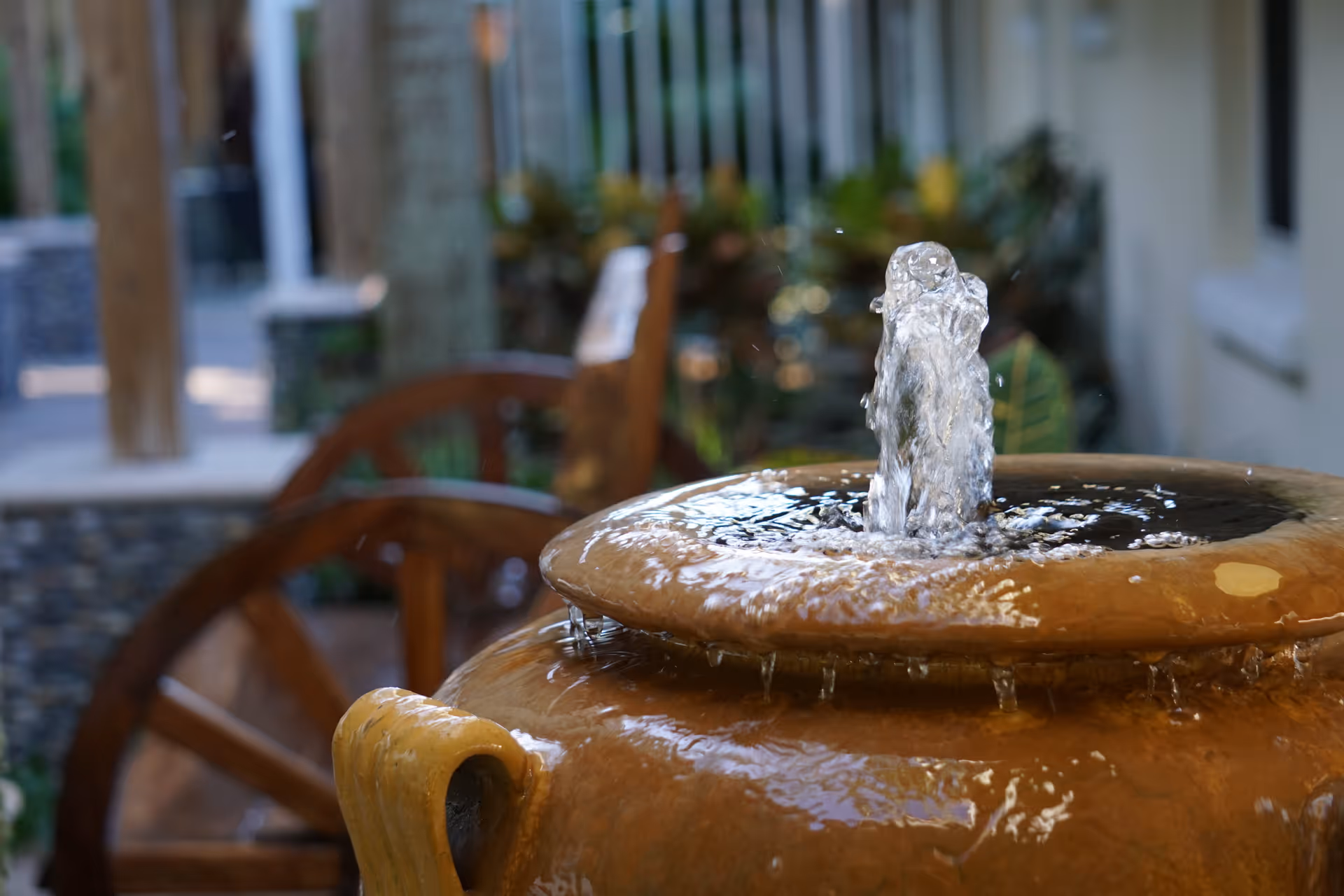 Close-up of a water fountain with water bubbling up from a large ceramic pot, with blurred wooden chairs and greenery in the background.