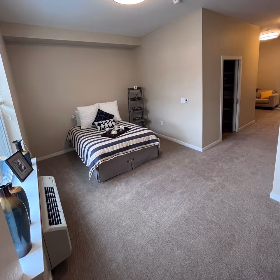 A bedroom with a single bed covered in a striped blanket and several pillows. Next to the bed is a small metal shelf with decorative items. The room has beige walls and carpeted floor. A window with a windowsill holding vases and a framed photo is on the left side. In the background, an open doorway leads to another room with a beige couch and a yellow pillow.