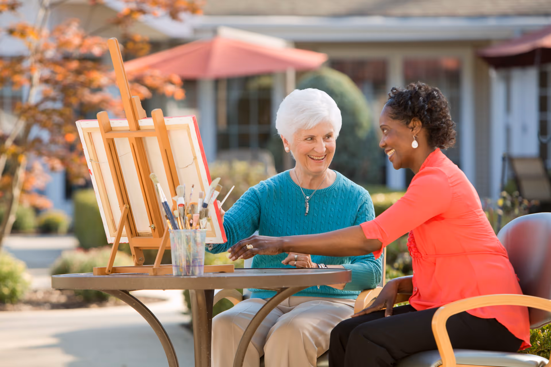An elderly white woman and a middle-aged Black woman sitting outside at a round table with an easel and painting supplies, smiling and engaging in a painting activity. The background shows a building with windows and umbrellas, suggesting a pleasant outdoor setting at a senior living facility.