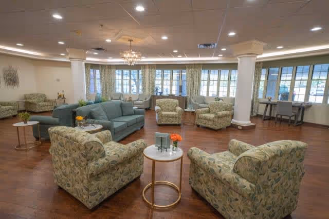 A spacious and well-lit common area with multiple upholstered armchairs and sofas arranged around small round tables. The room features large windows with curtains, wooden flooring, and a chandelier hanging from the ceiling, creating a comfortable and inviting atmosphere.