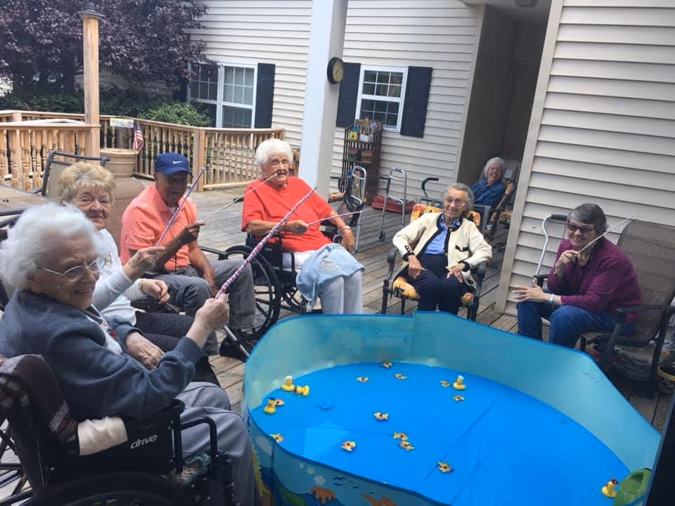A group of elderly people sitting outdoors on a wooden deck, engaging in a fishing game with toy fishing rods and floating rubber ducks in a small blue plastic pool. Some are seated in wheelchairs and others in chairs, enjoying the activity together.