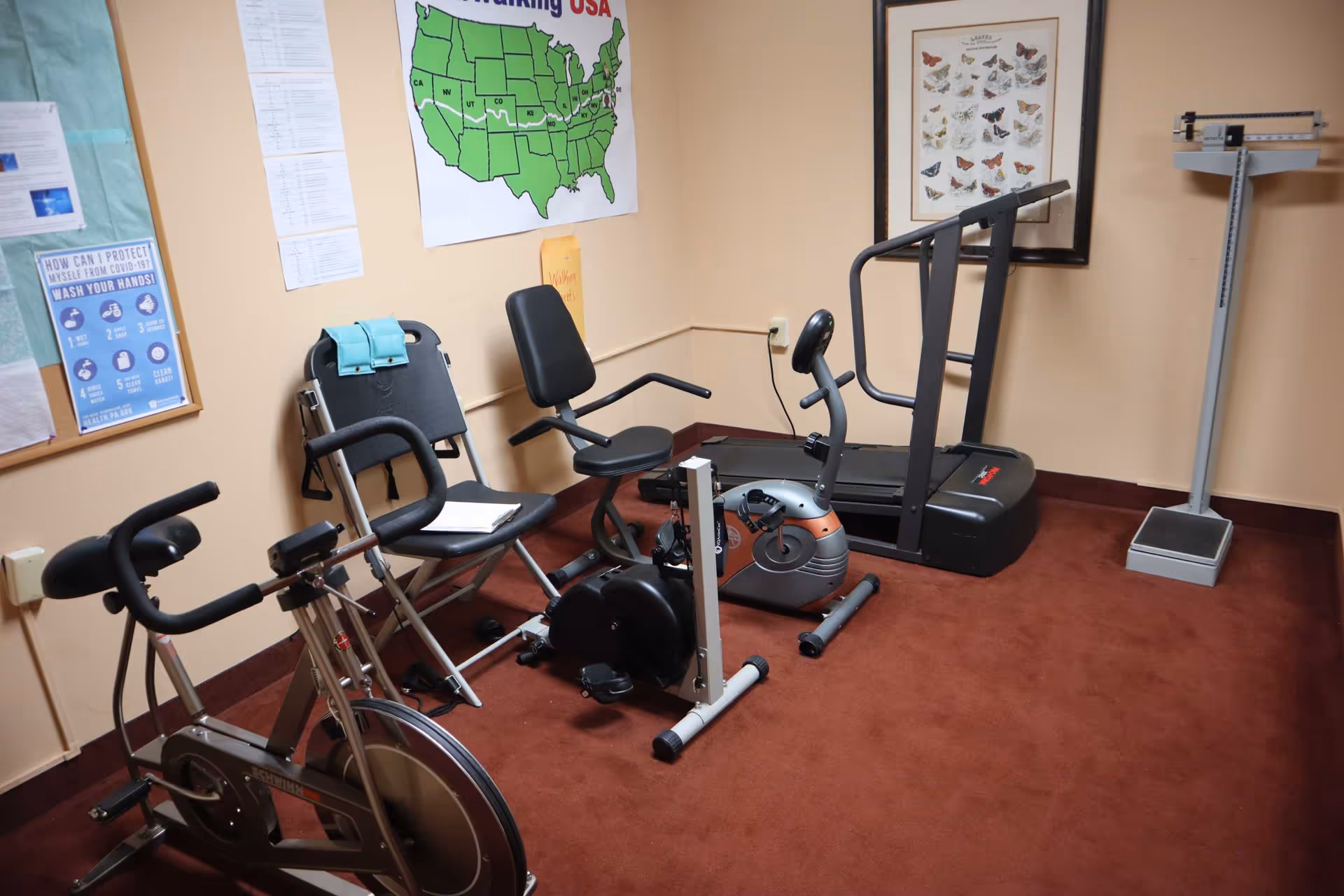 A small exercise room with various fitness equipment including a stationary bike, a recumbent bike, a treadmill, and a scale. The room has beige walls with posters and a map of the United States on the wall. The floor is covered with reddish-brown carpet.