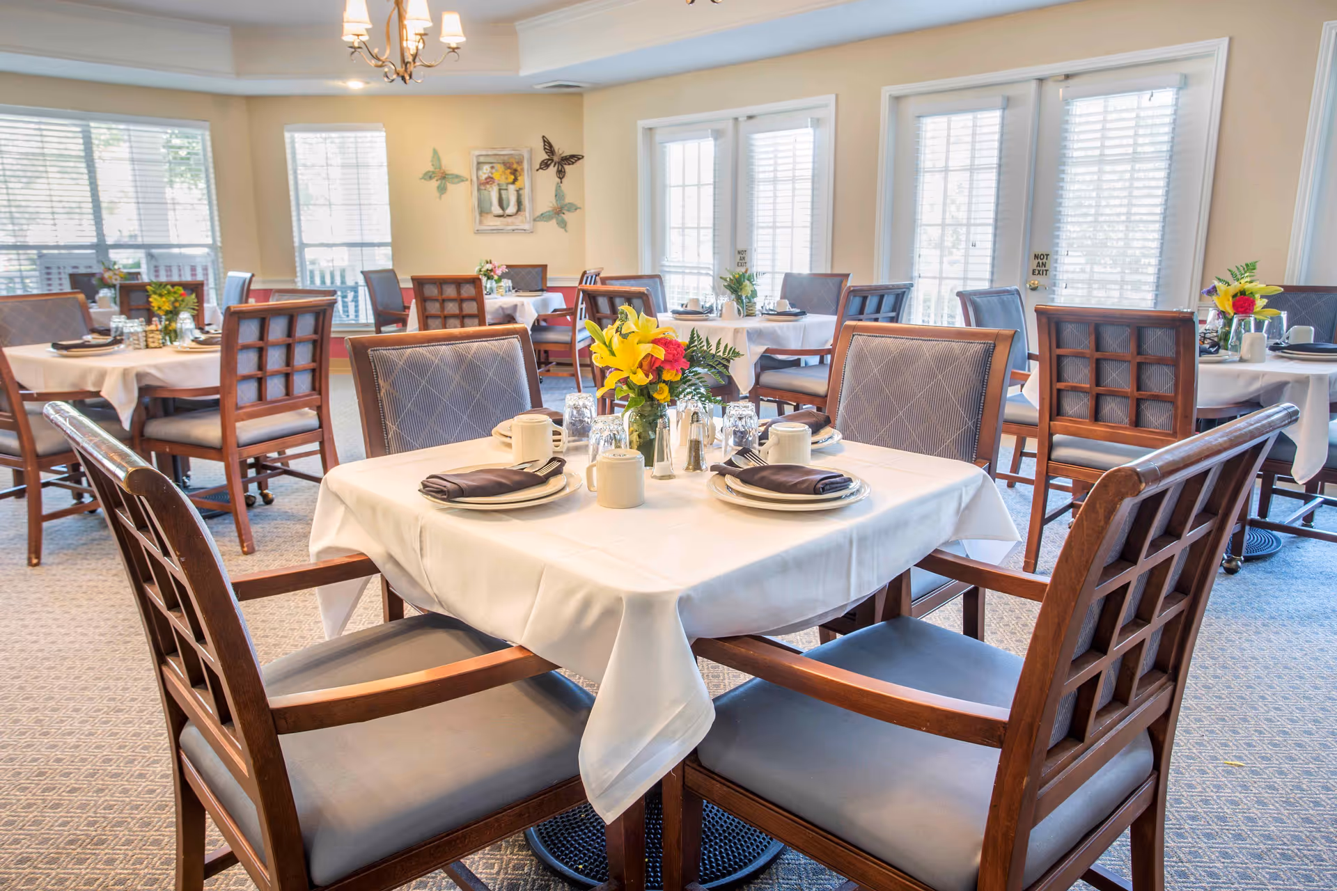 Bright dining room with tables set with white linens, wooden chairs, and floral centerpieces.