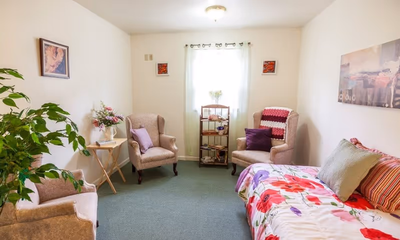 A cozy room with three beige armchairs, a small wooden side table with a vase of flowers, a small shelving unit in front of a window with sheer white curtains, and a bed with a colorful floral bedspread and multiple pillows. The walls are decorated with framed artwork and there is a green carpet on the floor.