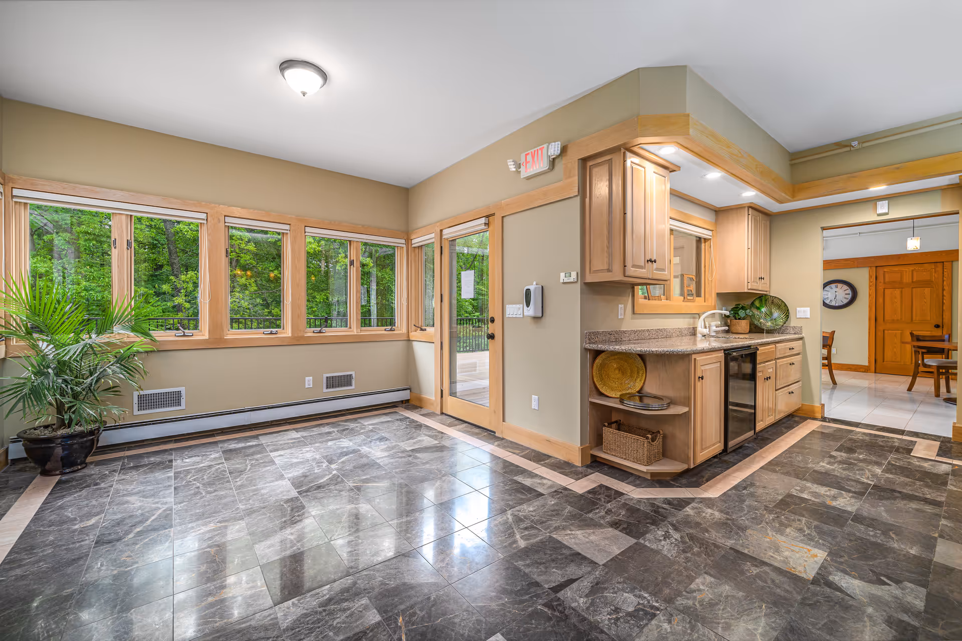 Interior view of a room with large windows showing green trees outside, a tiled floor with a decorative border, a small kitchen area with wooden cabinets, a countertop, and a sink. There is a potted plant in the corner and a door leading outside. Another room with a wooden door and a clock on the wall is visible in the background.