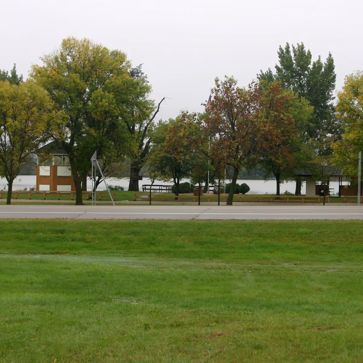 A grassy area with a road in the middle ground and a row of trees beyond it. Behind the trees, there is a body of water and some park benches and small structures visible.