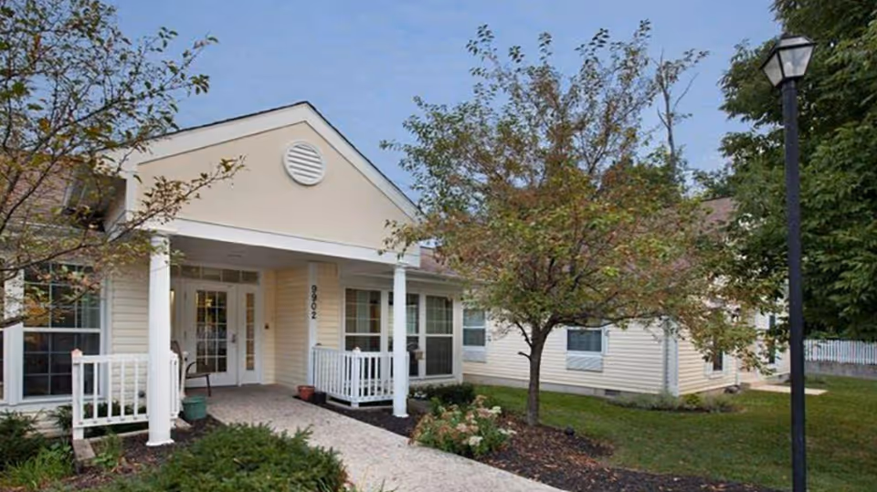 Exterior view of a single-story residential building with beige siding, white trim, and a covered porch supported by white columns. There are trees and shrubs in the landscaped front yard, and a black lamppost is visible on the right side of the image under a blue sky.