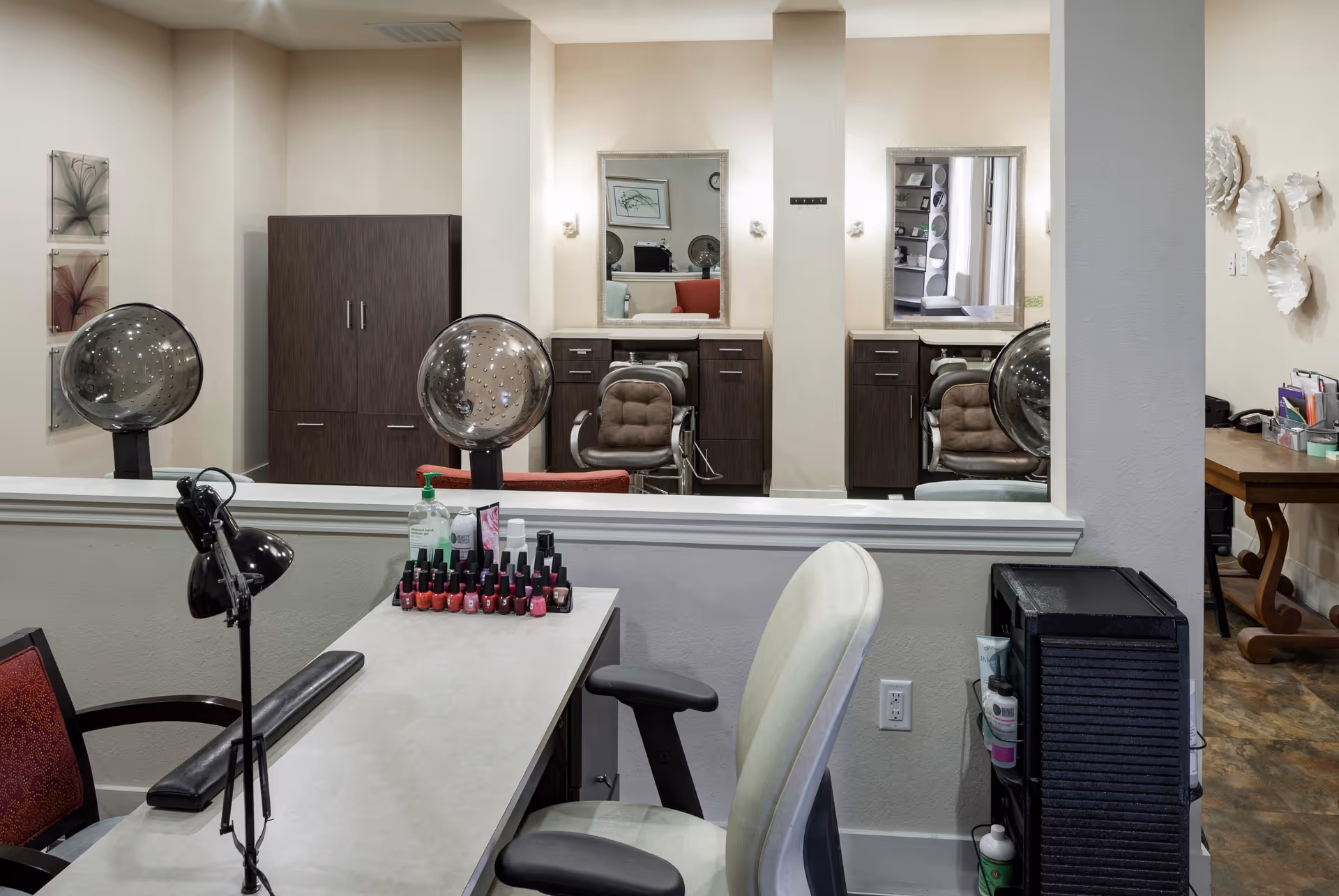 Interior salon area with a manicure table, nail polish display, chairs and hair-drying stations reflected in mirrors.