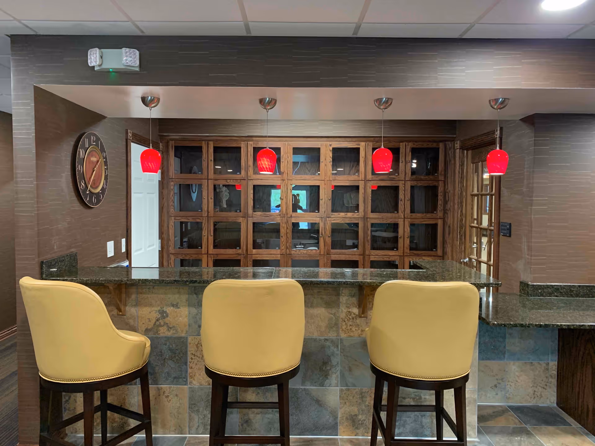 Interior view of a reception or common area with a granite countertop bar and three beige cushioned bar stools. Behind the counter is a wooden cabinet with glass doors and four red pendant lights hanging from the ceiling. A round wall clock is mounted on the left wall.