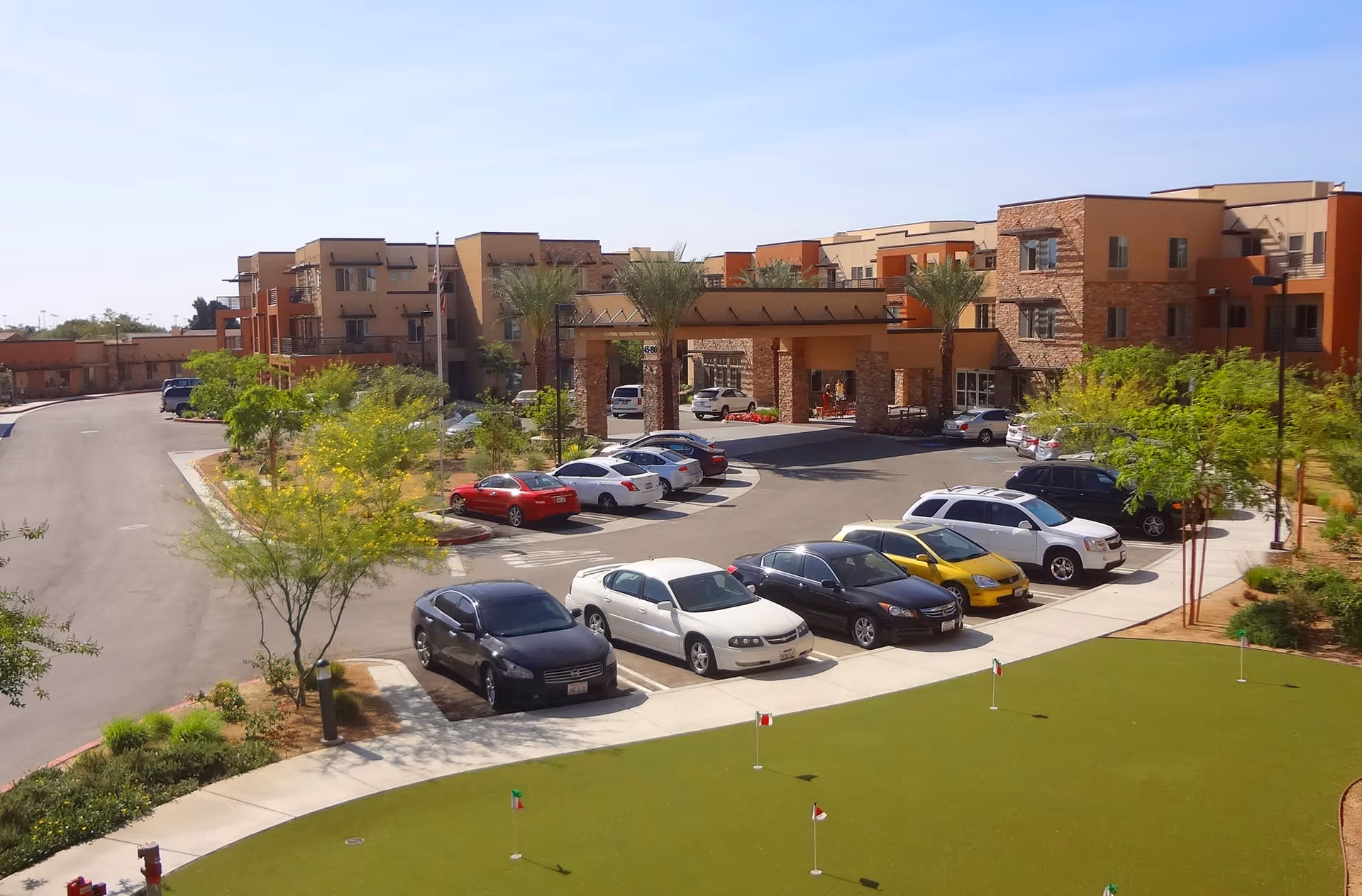 Exterior view of The Palms At La Quinta Gracious Retirement Living facility showing a parking lot with several cars, landscaped greenery, palm trees, and a putting green in the foreground. The multi-story building with stone and stucco facade is visible in the background under a clear blue sky.