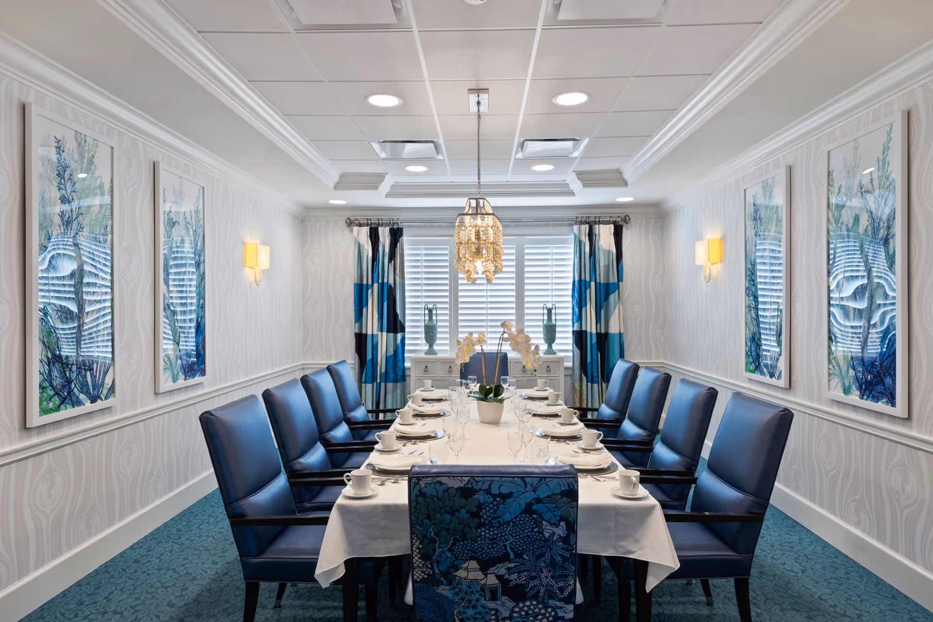 A formal dining room with a long rectangular table covered with a white tablecloth, set with white plates, cups, and glassware. The table is surrounded by blue upholstered chairs, with a patterned chair at the near end. The walls are decorated with framed artwork featuring aquatic themes, and there are blue and white curtains on the window at the far end. A chandelier hangs above the table, and the room has a blue carpet and white patterned wallpaper.