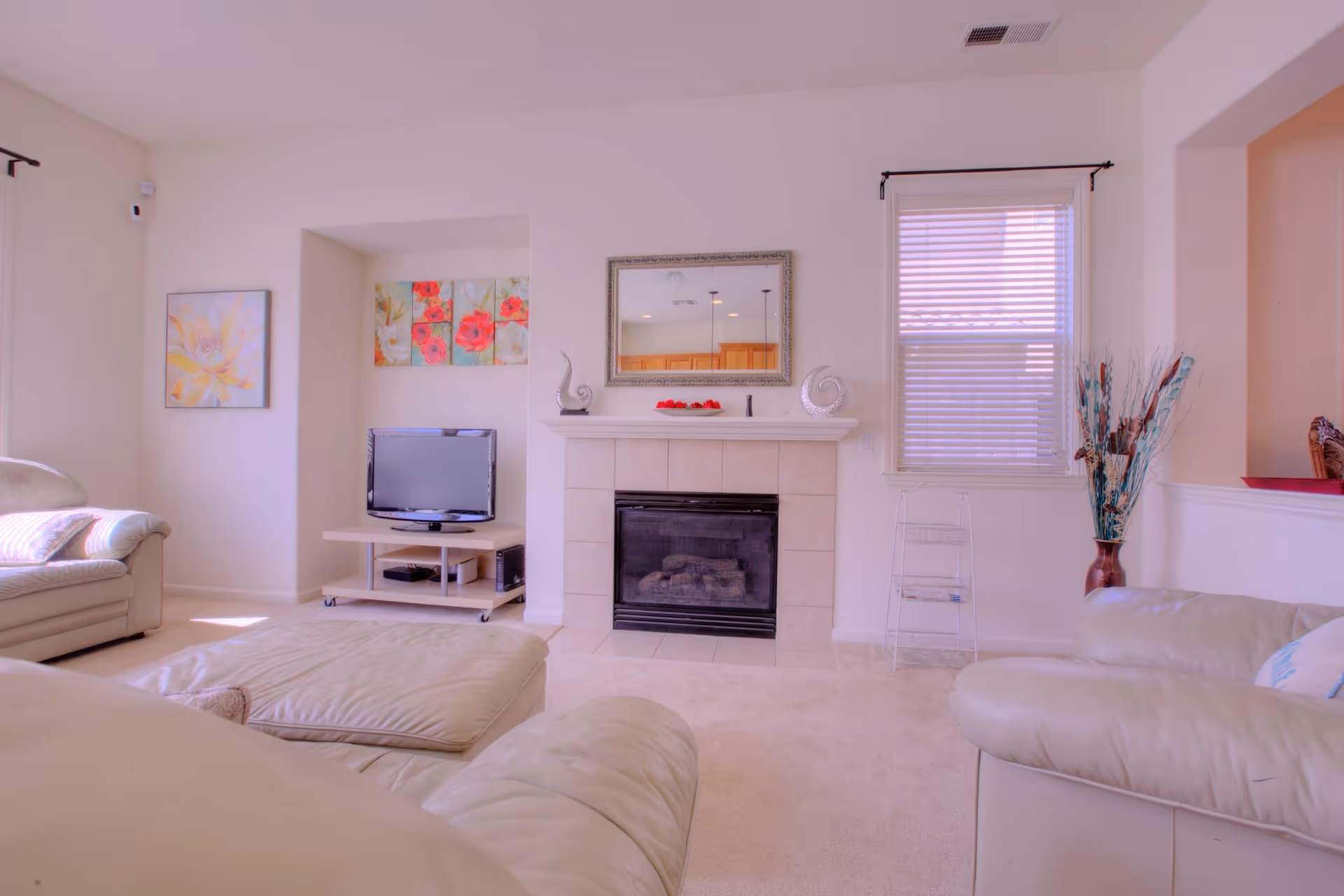 A cozy living room with cream-colored leather sofas and a matching ottoman. There is a fireplace with a decorative mirror above it, flanked by two ornamental sculptures. A flat-screen TV sits on a small stand in a recessed wall area, with colorful floral artwork above it. A window with blinds is on the right side, next to a tall vase with decorative branches. The room has light-colored walls and carpeted flooring.