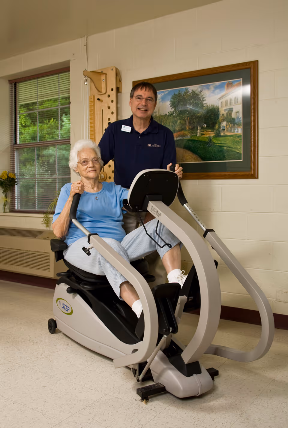 An elderly woman using a NuStep exercise machine indoors, assisted by a smiling male staff member standing behind her. The room has a window with blinds, a framed painting on the wall, and light-colored walls and floor.