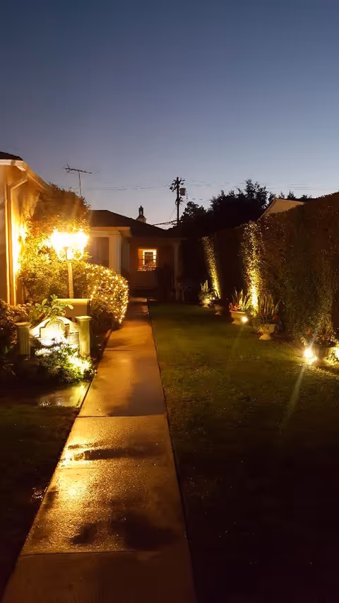 A dimly lit nighttime pathway through a garden courtyard with hedges, lawn, and exterior lights leading to a house.